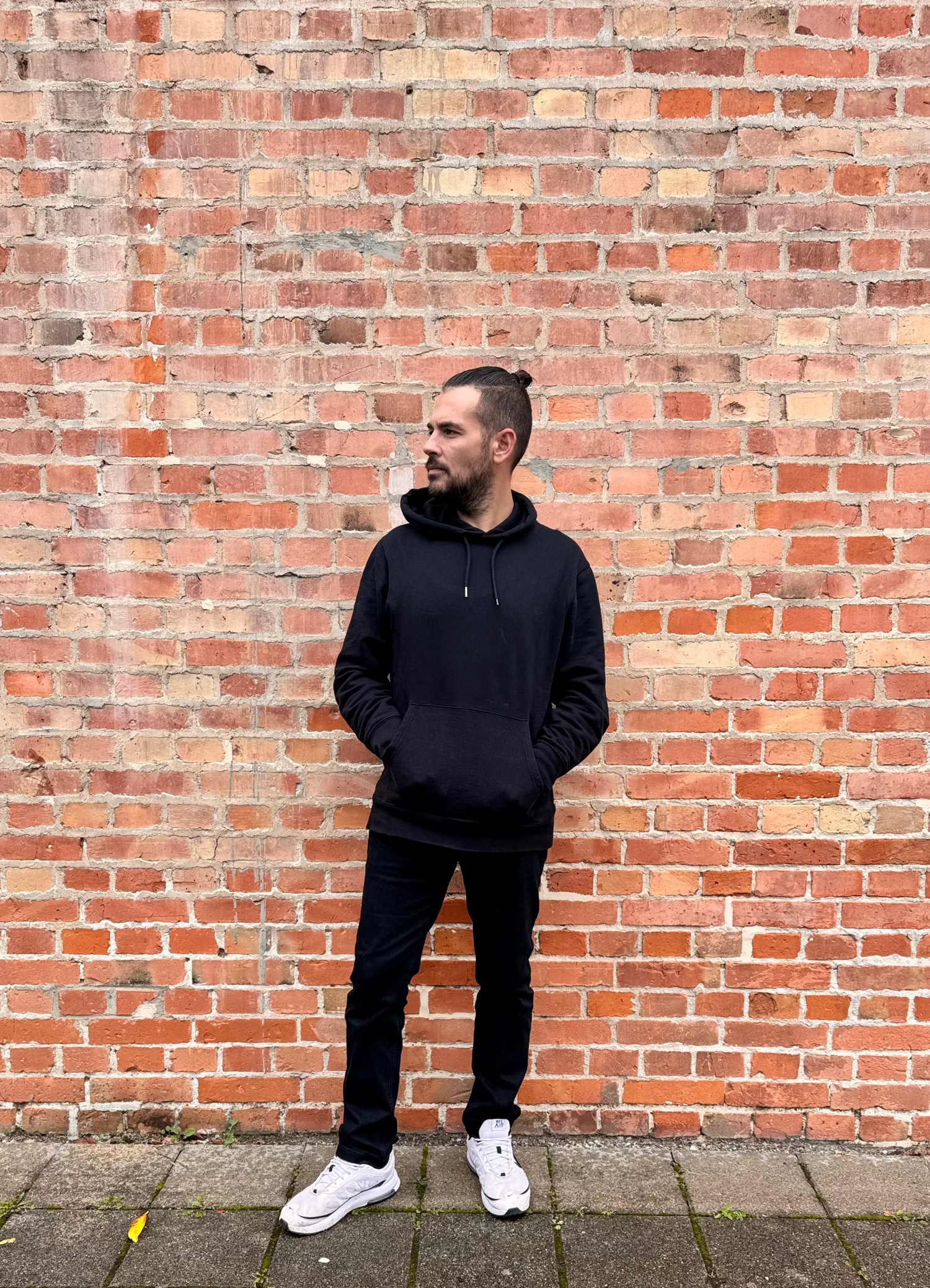 Mann in schwarzem Hoodie und Hose posiert vor Backsteinwand
